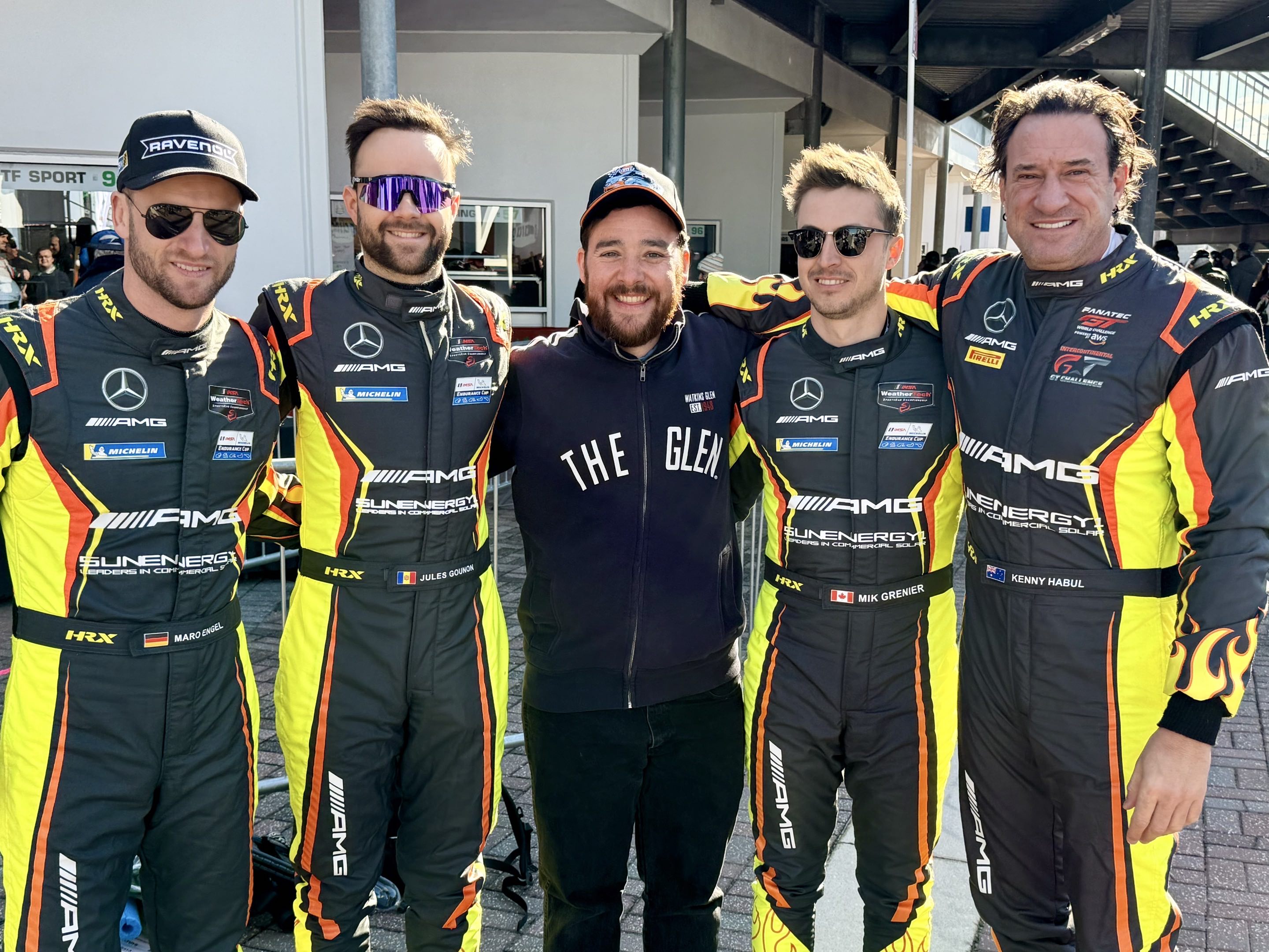 Four cool men in race suits flanking a very happy fan.